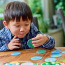 A child in a plaid shirt sits at a table, focused and smiling slightly, while playing with colourful, round game tokens. Bright, sunny atmosphere.