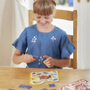 A smiling young girl in a blue dress plays a matching game at a wooden table. She holds a small card piece, showing focus and enjoyment.