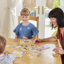 A woman and two children sit at a table playing a board game in a bright room. They are smiling and engaged, creating a joyful, playful atmosphere.