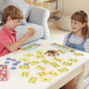 Two children play a board game at a white table, smiling and engaged. The game features yellow cards with paw prints and a circular spinner.