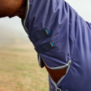 Partial view of a horse wearing a blue protective blanket with two adjustable straps. The background is a blurred, grassy field, creating a calm atmosphere.