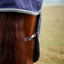 Close-up of a horse's brown tail covered with a navy blanket, secured with a metal clasp. The background is a blurry open field.