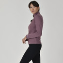 Woman in a mauve zip-up jacket and black leggings looks to the side against a light grey background, conveying a calm and confident tone.