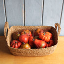 A rustic woven basket containing a collection of artificial autumn pumpkins and gourds in warm seasonal colours, set against a wooden background.