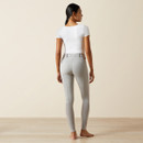 A person with dark hair in a low ponytail stands barefoot on a wooden floor, wearing a white short-sleeve shirt and gray fitted pants, viewed from the back.