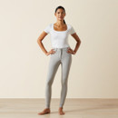 A confident woman stands on a wooden floor against a neutral background, wearing a white top and grey leggings, hands on hips, smiling slightly.