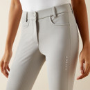 A close-up of a person wearing light gray riding breeches with "Ariat" on the leg. The fabric appears stretchy and the fit snug, conveying comfort.