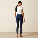 A woman with long dark hair, in a white shirt and navy riding pants, stands barefoot on a wooden floor, facing away in a neutral-toned room.