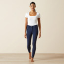 A woman in a white top and navy riding pants stands barefoot on a wooden floor against a neutral background. She appears confident and poised.