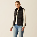 A person wearing a black puffer vest over a beige jumper stands confidently against a light background, paired with blue jeans, conveying casual style.