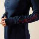 A person wearing a dark blue jacket with red "ARIAT" lettering on the sleeve. Their hands are clasped softly, conveying a relaxed tone.