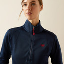 Woman wearing a navy high-collar pullover with a red logo and zip against a neutral background. She appears calm and confident.