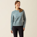 Smiling woman wearing a blue sweatshirt with "ARIAT" text and black leggings. She stands against a neutral background, exuding a casual and relaxed vibe.