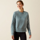 Smiling woman wearing a blue "Ariat" sweatshirt stands against a neutral background. Her relaxed pose and natural lighting create a warm, casual vibe.