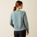 A woman with brown hair in a ponytail, wearing a blue long-sleeve top and black patterned leggings, stands facing away against a beige background.