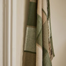 A wool blanket with a green and beige checked pattern hanging against a cream-coloured wall.