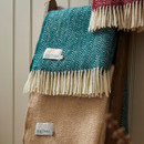 A teal wool blanket with a herringbone pattern and cream fringe folded on a ladder with beige and red blankets.