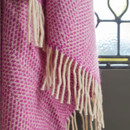 A pink wool blanket with a beehive pattern and cream fringe hanging near a window.