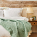 A mint green wool blanket with a waffle texture and cream fringe draped across a bed.
