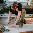 A person wearing protective gear, including goggles and earmuffs, uses a power saw to cut a concrete block outdoors. The setting is a construction site.