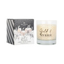 Cream candle in a clear glass holder with "Gold & Myrrh" written on the front, displayed beside its black and white gift box illustrated with a snowy village scene. The packaging highlights amber, fruity, and warm scents.