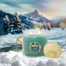 A green Yankee Candle burning with two wicks, labelled “Holiday Winterfest”. It is placed in snow with a slice of lemon and pine sprigs beside it, set against a snowy mountain landscape at sunset.