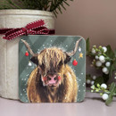A festive square coaster featuring an illustrated Highland cow with long shaggy hair, decorated with Christmas baubles and a gold star hanging from its horns, set against a green background with falling snow.