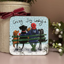 A square coaster featuring an illustration of a woman in a red bobble hat and blue coat sitting on a green bench with four dogs, watching the snowfall. The text above reads “Crazy Dog Lady” in a handwritten style.