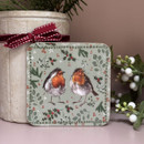 A square coaster decorated with an illustration of two robins facing each other. The birds have red breasts and fluffy feathers, set against a sage green background patterned with holly, berries, fir branches and white stars for a festive design.