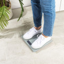 Person in blue jeans and white sneakers standing on a digital scale. The setting is a tiled floor with a potted plant nearby, creating a calm, natural tone.