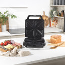 A black sandwich maker sits open on a white kitchen counter. Nearby are a loaf of bread, sliced ham, cheese, tomatoes, and an onion on a wooden board.