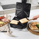 A sandwich maker with toasted sandwiches is on a worktop. Nearby, tomatoes, ham, and cheese are on a cutting board. A hand serves the sandwiches, creating a cosy kitchen scene.