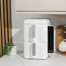 Dual white air fryer with silver handles on a kitchen counter. Decor includes a small potted plant, wooden board, and white jug. Bright, modern setting.
