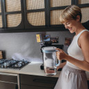 Smiling woman holding a water filter pitcher in a modern kitchen. She wears a white tank top and beige skirt. The ambience is warm and inviting.