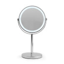 A round, illuminated tabletop mirror on a sleek silver stand, against a white background. The minimalist design conveys elegance and simplicity.