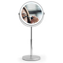 A round magnifying mirror on a stand showing a close-up of a person applying mascara. The scene feels focused and personal, with a clean background.
