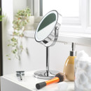 A vanity setup with a round mirror, an orange makeup brush, a razor, and a yellow soap dispenser on a white surface. A plant and cotton balls add a fresh touch.