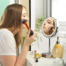 A woman applies makeup with an orange brush while looking into a round mirror. Nearby are makeup tools, a soap dispenser, and a potted plant, conveying a calm, focused tone.