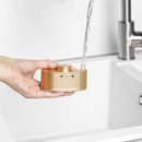 A hand holds a small, golden soap dispenser under a running tap in a white sink. The dispenser has lock symbols, indicating open and closed positions.