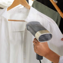 A hand holds a garment steamer, smoothing a white shirt on a hanger, with a green garment in the background. The scene conveys a clean, tidy feel.