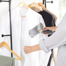 A person is using a handheld steamer on a white shirt hanging on a rack. Other clothes, including a dark shirt and beige jumper, are visible. The scene is calm and tidy.