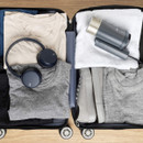 Open suitcase with neatly packed clothes, including beige and grey shirts, grey shoes, a white towel, blue headphones, and a grey steamer. Wooden floor.