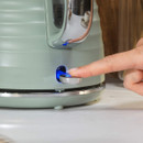 A finger presses a blue-lit button on a pale green electric kettle with a chrome handle. The scene suggests a modern, functional kitchen setting.