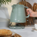 A hand lifts a sleek, mint green electric kettle off its base in a kitchen setting. Nearby are toasted bread, a white mug, and wooden utensils. The scene feels warm and inviting.
