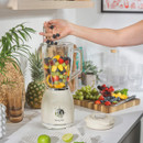 A hand adds blueberries to a blender filled with various fruits, surrounded by fresh ingredients and kitchen décor.