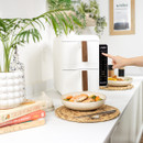 A kitchen counter with a modern white air fryer and a hand pressing its digital display. Plates of food, cookbooks, and plants create a fresh, inviting space.