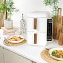 Modern kitchen counter displaying a sleek white air fryer with wooden handles and digital controls. Nearby, dishes with salmon and onion rings are served, set against a backdrop of cookbooks, plants, and elegant decor.