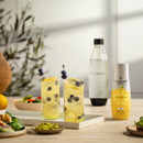 Tall glasses of fizzy lemonade with lemon slices and blueberries on a tray. Nearby are a Sodastream bottle and syrup, with a bowl of salad and guacamole. Bright, refreshing vibe.