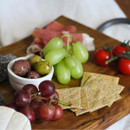 Green grapes, red grapes, tomatoes on the vine, olives, and crackers displayed on a wooden board.