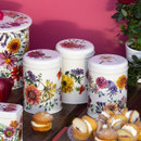 Set of Emma Bridgewater Garden Flowers storage caddies with colourful floral patterns, displayed on a wooden table alongside cakes and fresh flowers, against a pink background.